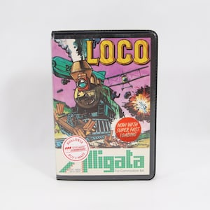 Puede incluir: Cartera negra con una colorida ilustración de un tren, un avión y explosiones, con la palabra "LOCO" en amarillo. También se ve el texto "Now with Super Fast Loading". La cartera es para Commodore 64.