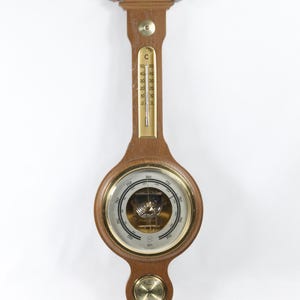 Puede incluir: Una estación meteorológica de pared de madera con un barómetro, un termómetro y un higrómetro. El barómetro tiene una cara redonda con un borde dorado y una esfera blanca con marcas negras. El termómetro tiene un tubo de vidrio largo y delgado con un líquido rojo en su interior. El higrómetro tiene una cara redonda pequeña con un borde dorado y una esfera blanca con marcas negras.