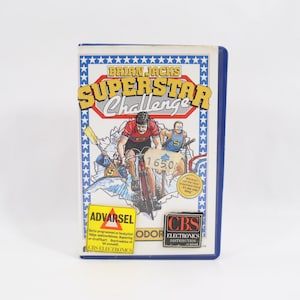 Puede incluir: Un cartucho de videojuego vintage para "Brian Jacks Superstar Challenge". La caja presenta un ciclista en rojo y negro, con el título en amarillo y rojo. La caja tiene un borde azul y un fondo blanco.