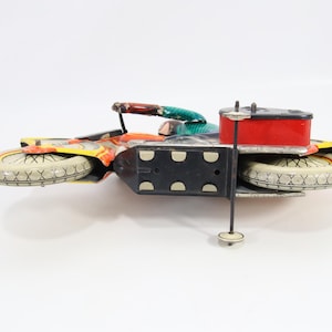 May include: Vintage toy motorcycle with a red fuel tank, black chassis, and white tires. The motorcycle has colorful accents of orange, yellow, and green. A classic collectible toy.