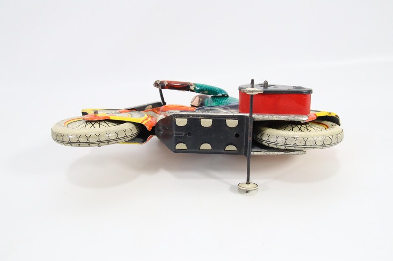 May include: Vintage toy motorcycle with a red fuel tank, black chassis, and white tires. The motorcycle has colorful accents of orange, yellow, and green. A classic collectible toy.