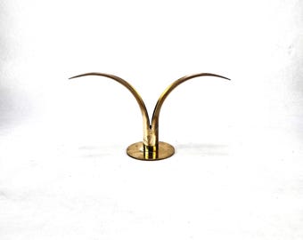 May include: A gold metal candle holder with two curved arms. The holder has a round base.