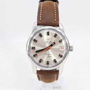 Puede incluir: Reloj automático Enicar Sherpa Date plateado con correa de cuero marrón. La esfera del reloj es plateada con agujas negras y naranjas, y una pantalla de fecha a las 3 en punto. El reloj tiene una esfera redonda y una correa de cuero marrón.