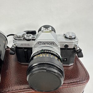 May include: A silver Canon AT-1 film camera with a black lens. The camera is sitting on a brown leather case.