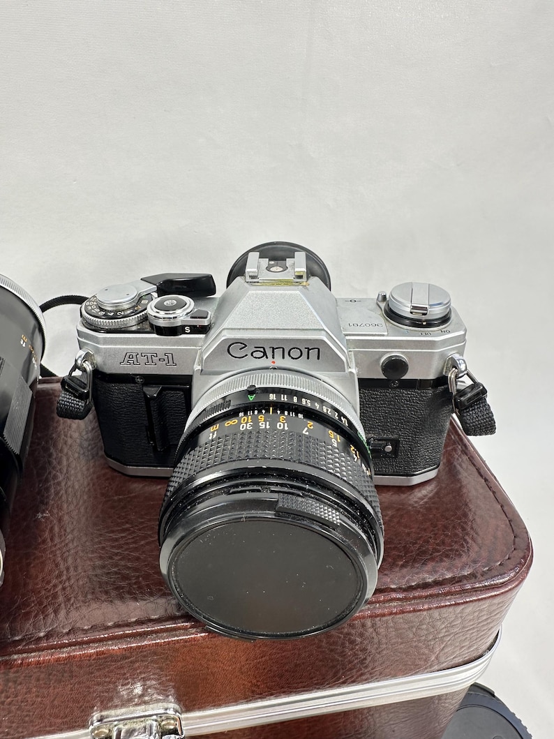 May include: A silver Canon AT-1 film camera with a black lens. The camera is sitting on a brown leather case.