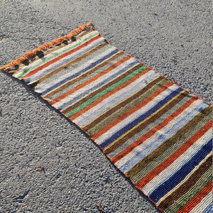 May include: A long, narrow, multi-colored striped rug with fringe on the ends. The rug is made of woven fabric and has a variety of colors, including red, orange, yellow, green, blue, and brown.