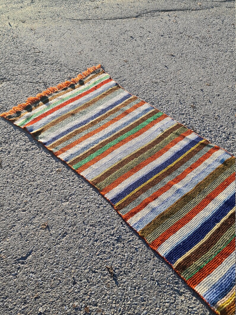 May include: A long, narrow, multi-colored striped rug with fringe on the ends. The rug is made of woven fabric and has a variety of colors, including red, orange, yellow, green, blue, and brown.