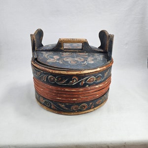 May include: A round wooden box with a lid, painted in black, red, and white with floral designs. The box has two wooden handles on the sides and a smaller handle on the lid.