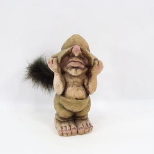 May include: A whimsical figurine of a troll, featuring a hooded outfit and a furry tail. The troll is depicted with its hands covering its eyes. The figurine is primarily beige and brown, with a touch of pink on the nose.