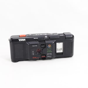 Puede incluir: Una cámara de película de 35 mm Toma OX-1C negra y roja con flash incorporado. La cámara tiene un botón rojo en la parte superior y una lente plateada.