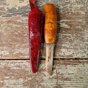 Puede incluir: Dos mangos de cuchillo de madera, uno con una vaina de cuero roja con tallas florales. La vaina est&aacute; cerrada y el cuchillo no es visible. El otro mango es de madera marr&oacute;n claro con una hoja de metal.