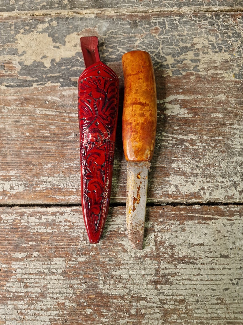 Puede incluir: Dos mangos de cuchillo de madera, uno con una vaina de cuero roja con tallas florales. La vaina est&aacute; cerrada y el cuchillo no es visible. El otro mango es de madera marr&oacute;n claro con una hoja de metal.