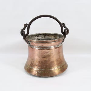 May include: An antique copper bucket with a dark metal handle. The bucket has a flared base and a hammered texture, showing signs of age and use. The handle is attached with decorative, curved brackets. A charming piece.
