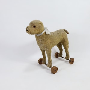 Puede incluir: Un perro de peluche marrón con una cinta blanca alrededor del cuello. El perro está sobre ruedas y tiene un aspecto vintage.