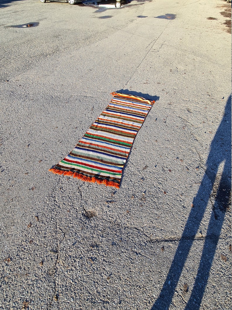 May include: A long, striped rug with orange, brown, white, blue, and green stripes. The rug has fringe on the ends and is lying on a gray asphalt surface.