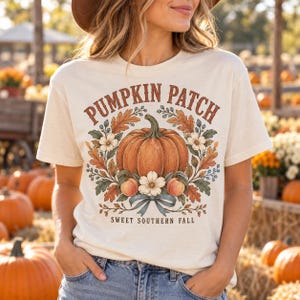 May include: A cream-colored short-sleeve t-shirt featuring a graphic of a large orange pumpkin surrounded by autumn leaves, small flowers, and berries. The text reads PUMPKIN PATCH at the top and SWEET SOUTHERN FALL at the bottom.