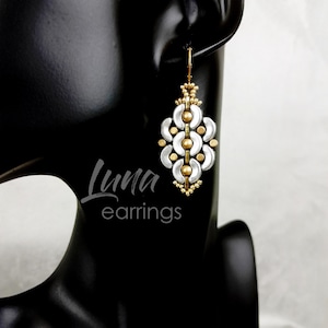 Puede incluir: Pendientes de cuentas blancas y doradas con un diseño geométrico. Los pendientes cuelgan de la oreja de un maniquí negro. El texto "Luna earrings" es visible debajo de los pendientes.