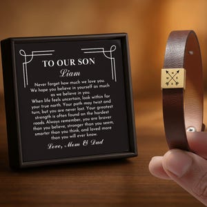 May include: A brown leather bracelet with a gold-colored compass charm. The bracelet is held next to a framed black card with the text "TO OUR SON Liam" and a heartfelt message from Mom & Dad.