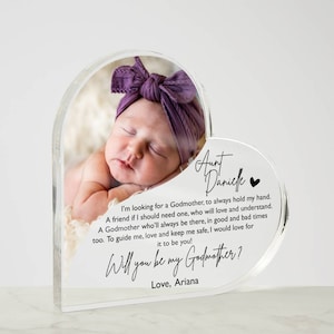 Peut inclure: Un cadre photo en forme de c&oelig;ur en acrylique transparent avec une photo d'un b&eacute;b&eacute; endormi. Le cadre contient un message demandant "Veux-tu &ecirc;tre ma marraine ?" et est sign&eacute; "Avec amour, Ariana".