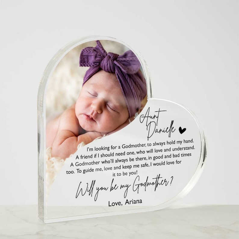 Peut inclure: Un cadre photo en forme de c&oelig;ur en acrylique transparent avec une photo d'un b&eacute;b&eacute; endormi. Le cadre contient un message demandant "Veux-tu &ecirc;tre ma marraine ?" et est sign&eacute; "Avec amour, Ariana".