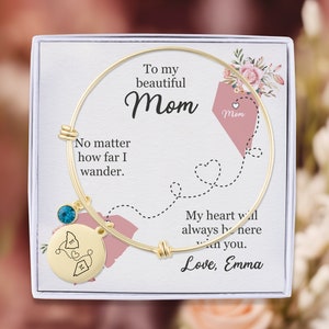 May include: A gold bangle bracelet with a charm that says "Mom" and a blue crystal. The bracelet is in a white box with a pink and white floral design and the text "To my beautiful Mom No matter how far I wander. My heart will always be here with you. Love, Emma".