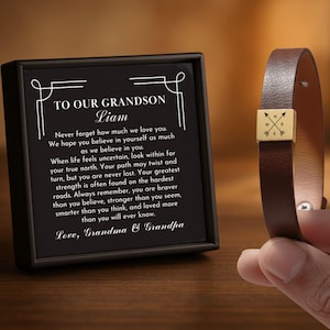 May include: A brown leather bracelet with a gold-colored square clasp featuring a compass rose design. Next to it is a framed black plaque with white text that reads "TO OUR GRANDSON Liam" and a heartfelt message from Grandma & Grandpa.