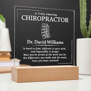 May include: A white acrylic plaque with a wooden base, featuring a personalized message for a chiropractor. The plaque reads "A Truly Amazing Chiropractor Dr. David Williams Is hard to find, difficult to part with, and impossible to forget. May you be proud of the work you do, the difference you make and the many lives you have touched!"