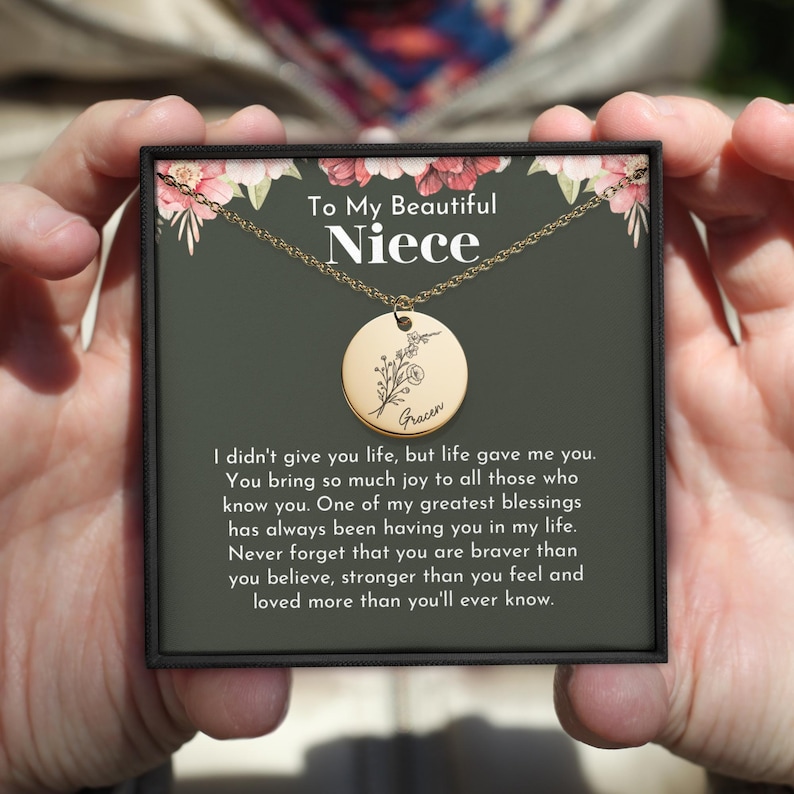 May include: A gold necklace with a round pendant featuring a floral design and the name "Fracen" engraved on it. The necklace is in a black box with a floral design and the text "To My Beautiful Niece".