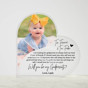 Peut inclure: Un cadre photo en acrylique en forme de c&oelig;ur avec une photo d'une petite fille portant un n&oelig;ud jaune. Le cadre porte le texte "Tis Fennie & Tia Fabi" et "Will you be my Godparents? Love, Layla".