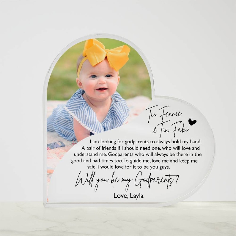 Peut inclure: Un cadre photo en acrylique en forme de c&oelig;ur avec une photo d'une petite fille portant un n&oelig;ud jaune. Le cadre porte le texte "Tis Fennie & Tia Fabi" et "Will you be my Godparents? Love, Layla".