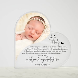 Peut inclure: Un cadre photo en acrylique blanc en forme de c&oelig;ur avec une photo d'une petite fille endormie. Le cadre porte un message qui dit "Oncle Nicky" et "Veux-tu &ecirc;tre mon parrain ? Avec amour, Ariana Jo".