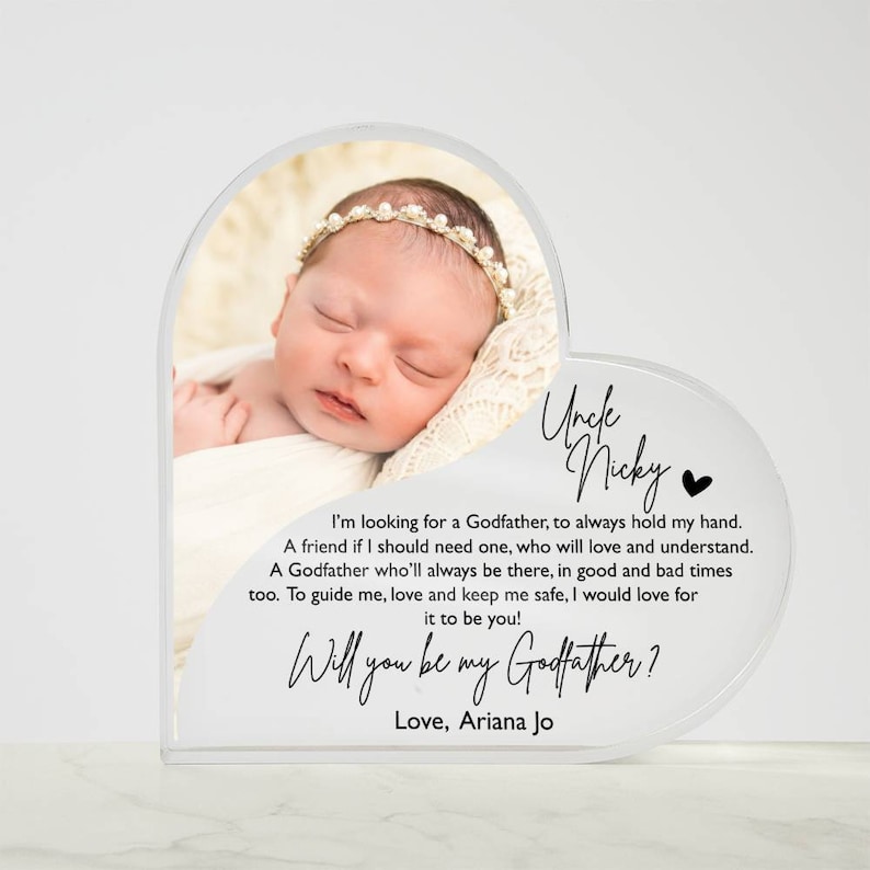 Peut inclure: Un cadre photo en acrylique blanc en forme de c&oelig;ur avec une photo d'une petite fille endormie. Le cadre porte un message qui dit "Oncle Nicky" et "Veux-tu &ecirc;tre mon parrain ? Avec amour, Ariana Jo".