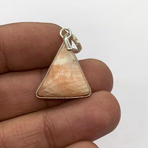 May include: A triangular pendant with a silver-colored frame and bail. The pendant features a peach-colored stone with a marbled appearance. The stone is set within a silver-colored metal frame. The bail is also silver-colored.