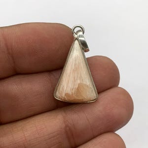May include: A triangular pendant with a silver-colored frame and bail. The pendant features a light peach-colored stone with a wood-grain pattern. The stone is set in a polished silver metal frame. The pendant is likely for a necklace.