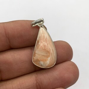 May include: A teardrop-shaped pendant with a silver-colored bail. The pendant features a peach-colored stone with a textured, layered appearance. The stone is encased in a silver-colored metal frame. The pendant is held in a hand.