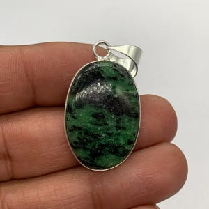 May include: An oval-shaped pendant featuring a polished green and black stone set in a smooth silver-toned metal frame. The pendant includes a silver-toned metal bail at the top, designed for attachment to a necklace chain or cord.