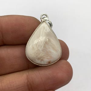 May include: A pear-shaped pendant with a silver bail. The pendant features a cream-colored stone with a unique, textured pattern. The stone is set in a silver bezel. The pendant is held in a person's hand.
