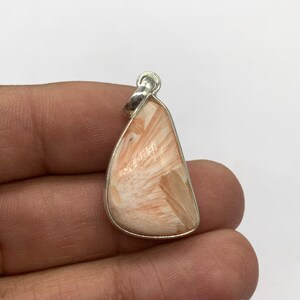 May include: A pendant with a silver-colored frame and a peach-colored stone. The stone has a unique pattern of light and dark lines radiating from the center. The pendant is held in a hand against a white background.