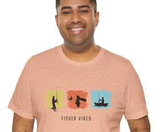 May include: Peach-coloured t-shirt featuring a graphic of three fishing scenes in colourful squares. The text "FISHER VIBES" is printed below the graphics. The shirt is worn by a person.