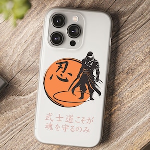 May include: A clear phone case featuring a black and orange graphic of a ninja warrior with Japanese text. The design includes a ninja figure, a rising sun, and Japanese characters. The case is on a wooden surface.