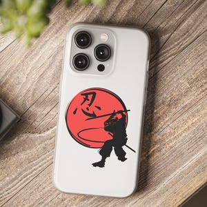 May include: Clear phone case with a black samurai warrior silhouette wielding a sword, set against a red sun. The Japanese character for "endurance" is above the warrior. The case is on a wooden surface.