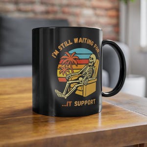 Op de afbeelding: Zwarte keramische mok met een humoristische afbeelding. Het ontwerp toont een skelet dat ontspant in een stoel onder een zonsondergang met palmbomen. De tekst luidt: "I'm still waiting for... IT SUPPORT."