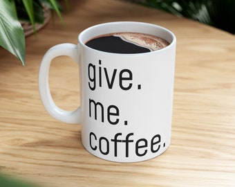 May include: White ceramic coffee mug with the words "give. me. coffee." in black, filled with dark coffee. The mug has a curved handle and sits on a light wood surface.