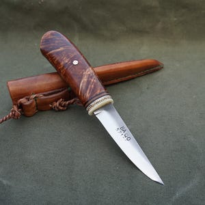 Op de afbeelding: Een handgemaakt mes met een houten handvat en een leren schede. Het mes heeft een zilverkleurig lemmet met de tekst "BAQO" en een serienummer. Het handvat is donkerbruin met een witte accent en een kleine witte stip.
