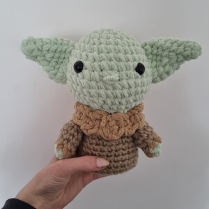 May include: A handmade crochet doll of a character with large ears and black eyes. The doll is primarily light green with a brown outfit. The doll is held in a person's hand against a white background.