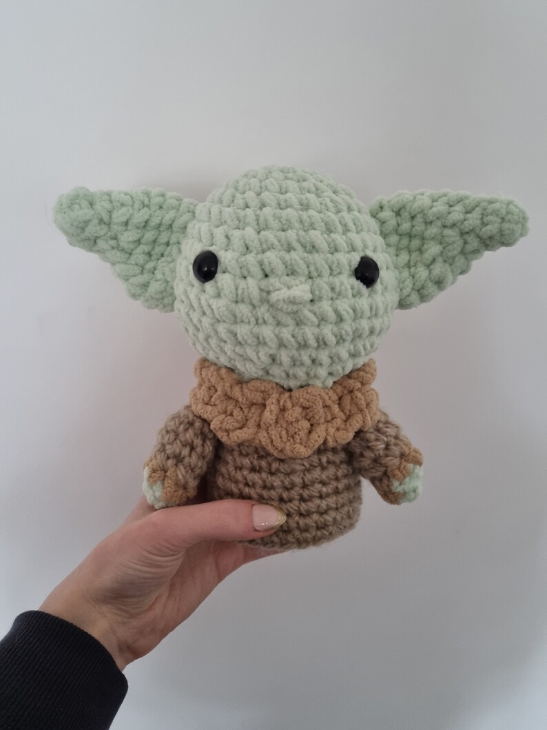 May include: A handmade crochet doll of a character with large ears and black eyes. The doll is primarily light green with a brown outfit. The doll is held in a person's hand against a white background.