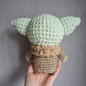 May include: A crocheted doll in the likeness of a character with large ears. The doll is primarily light green, with a tan neck and body. The doll is held in a hand against a gray background.