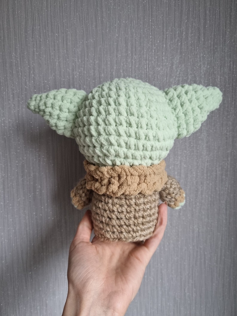 May include: A crocheted doll in the likeness of a character with large ears. The doll is primarily light green, with a tan neck and body. The doll is held in a hand against a gray background.