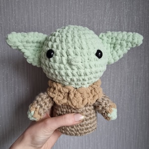 May include: A handmade crochet doll in the likeness of a popular character. The doll features large, pointed ears, black eyes, and a tan and green color scheme. The doll is held in a hand, showcasing the soft texture of the yarn.