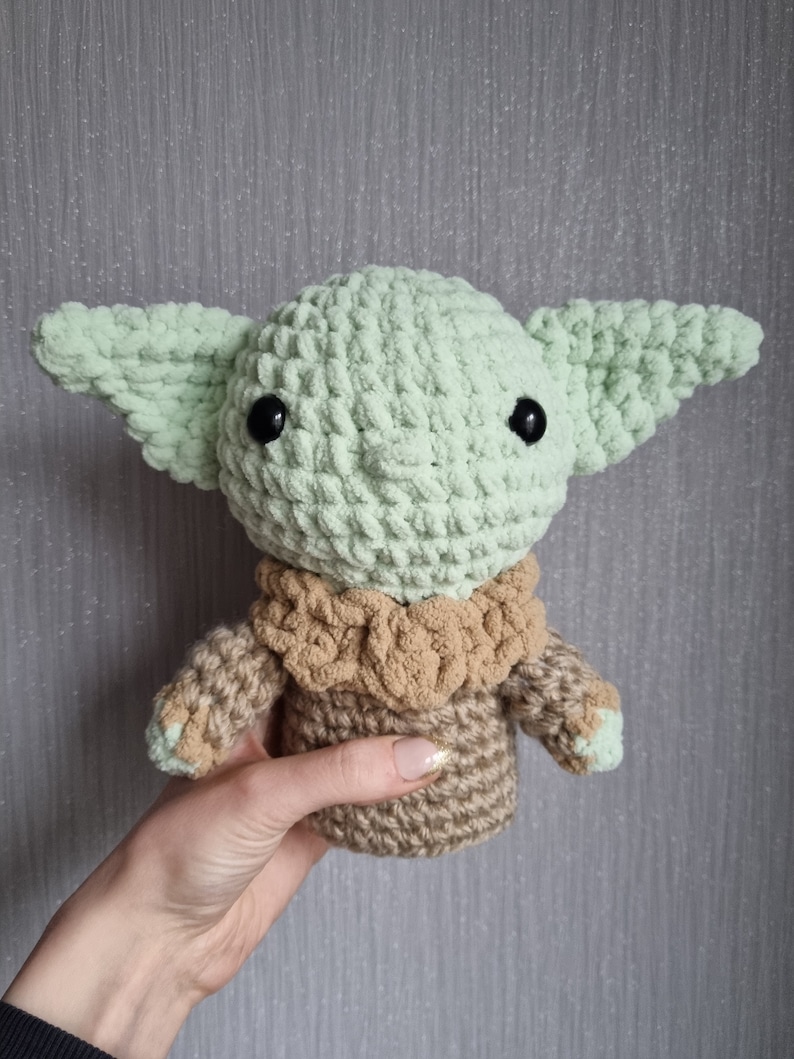May include: A handmade crochet doll in the likeness of a popular character. The doll features large, pointed ears, black eyes, and a tan and green color scheme. The doll is held in a hand, showcasing the soft texture of the yarn.